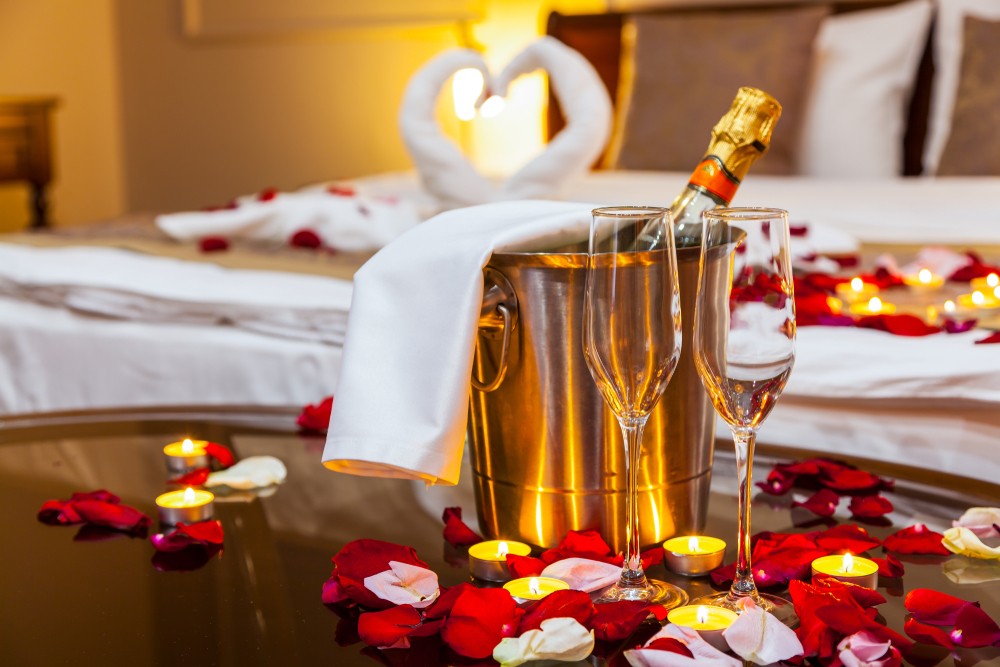 Romantic hotel room getaway, a table with a fruit plate and candles, in the background a bed decorated with swans of towels and rose petals