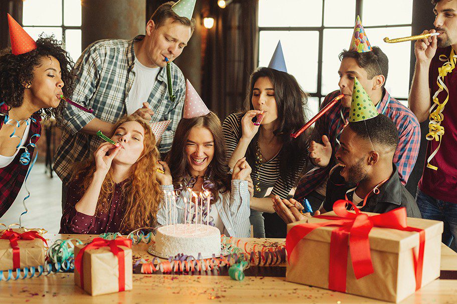 An image of a birthday party with adults surrounding a cake, wearing party hats.